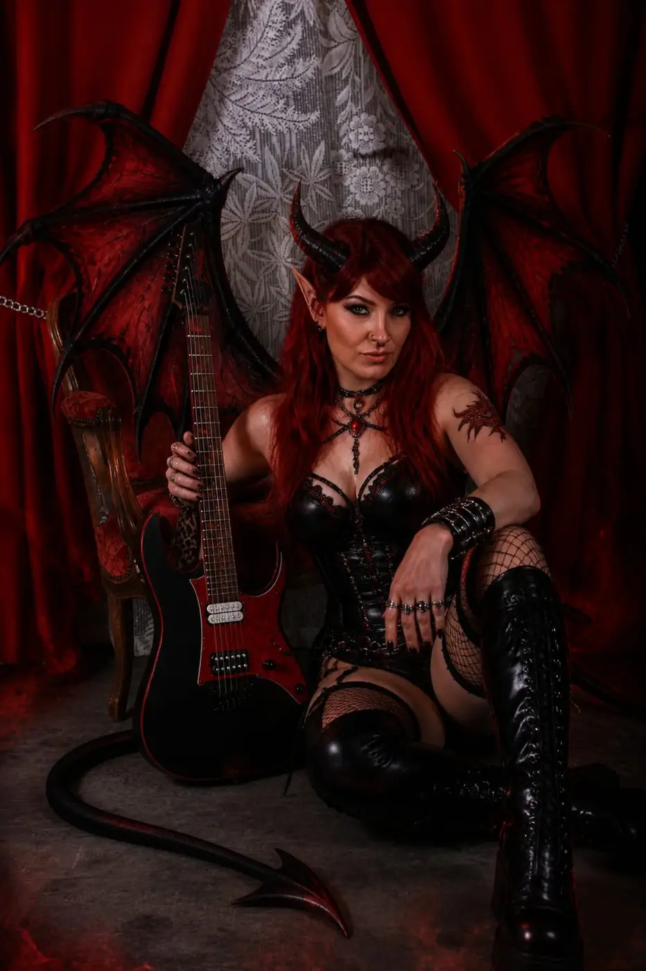 Portrait en pied d’une femme démon avec ailes, cornes et guitare, assise devant des rideaux rouges.