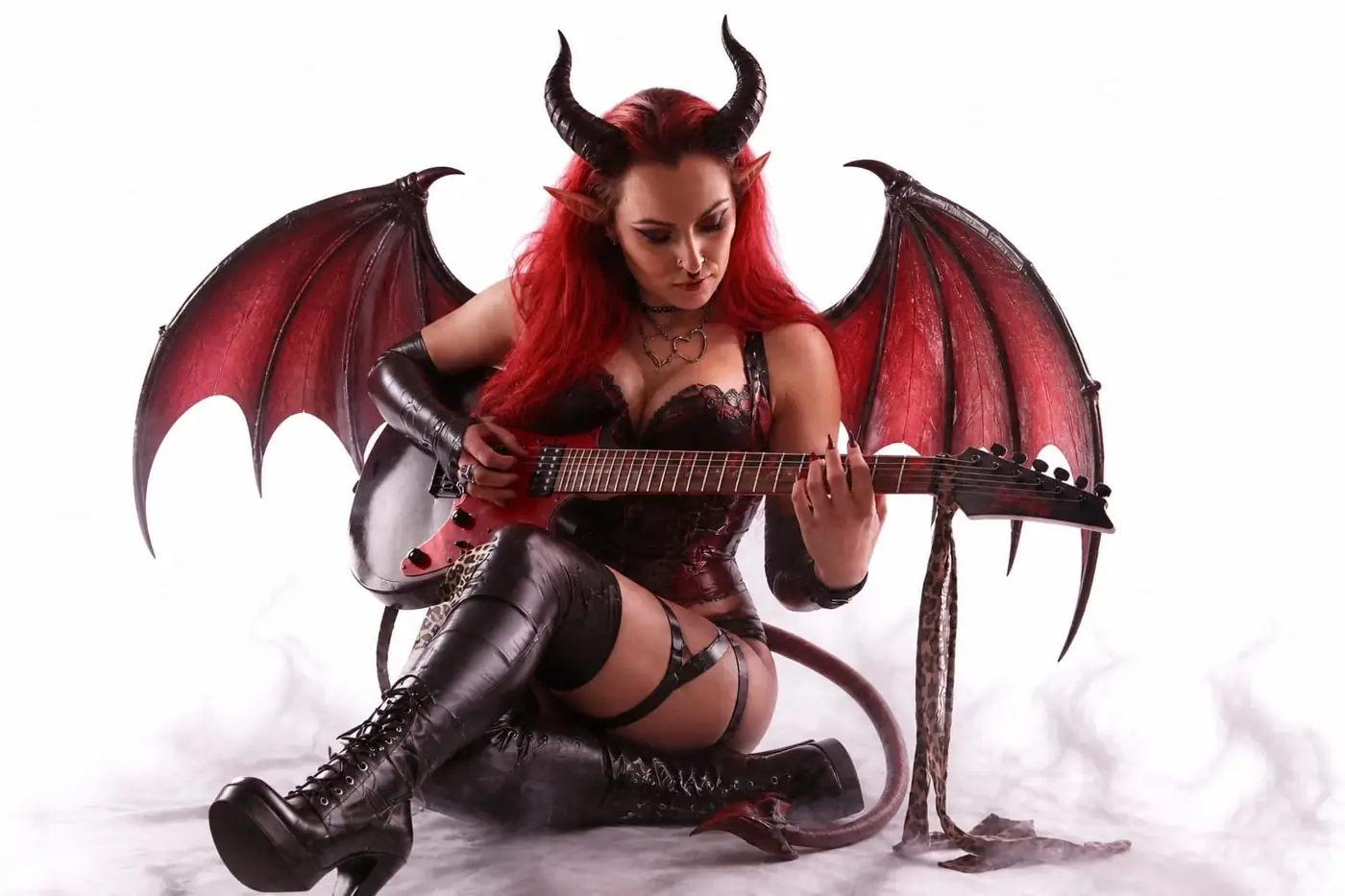 Création studio sur fond clair d’une femme démon jouant de la guitare avec ailes rouges et tenue dark fantasy.