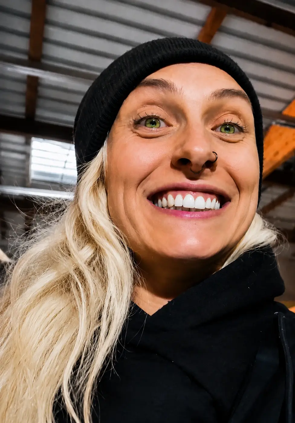 Portrait rapproché d’une femme blonde souriante portant un bonnet noir dans un intérieur industriel.